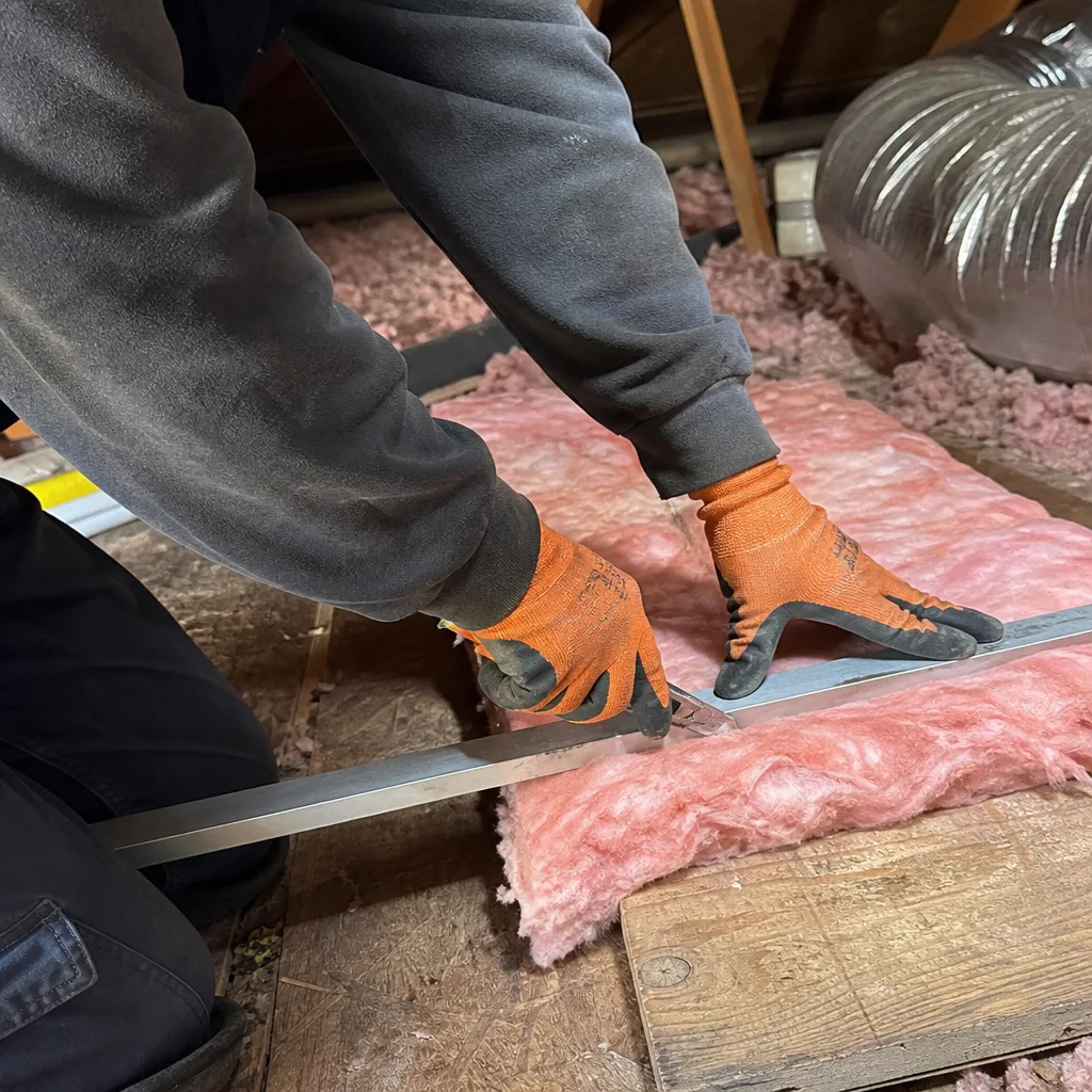Worker cutting pink fiberglass attic insulation batts with a straight edge
